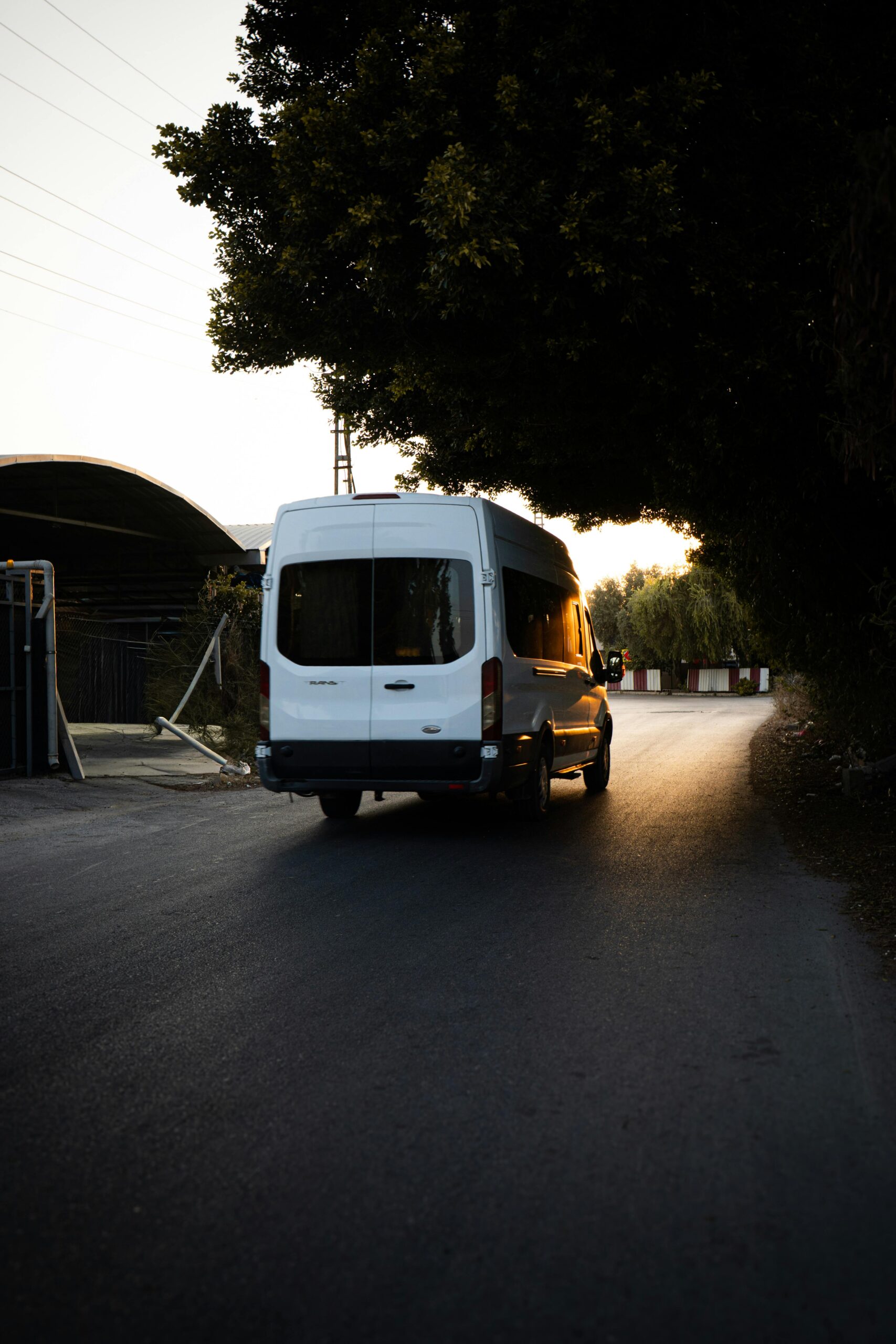 A white minivan drives on a quiet road under trees during a serene sunset.