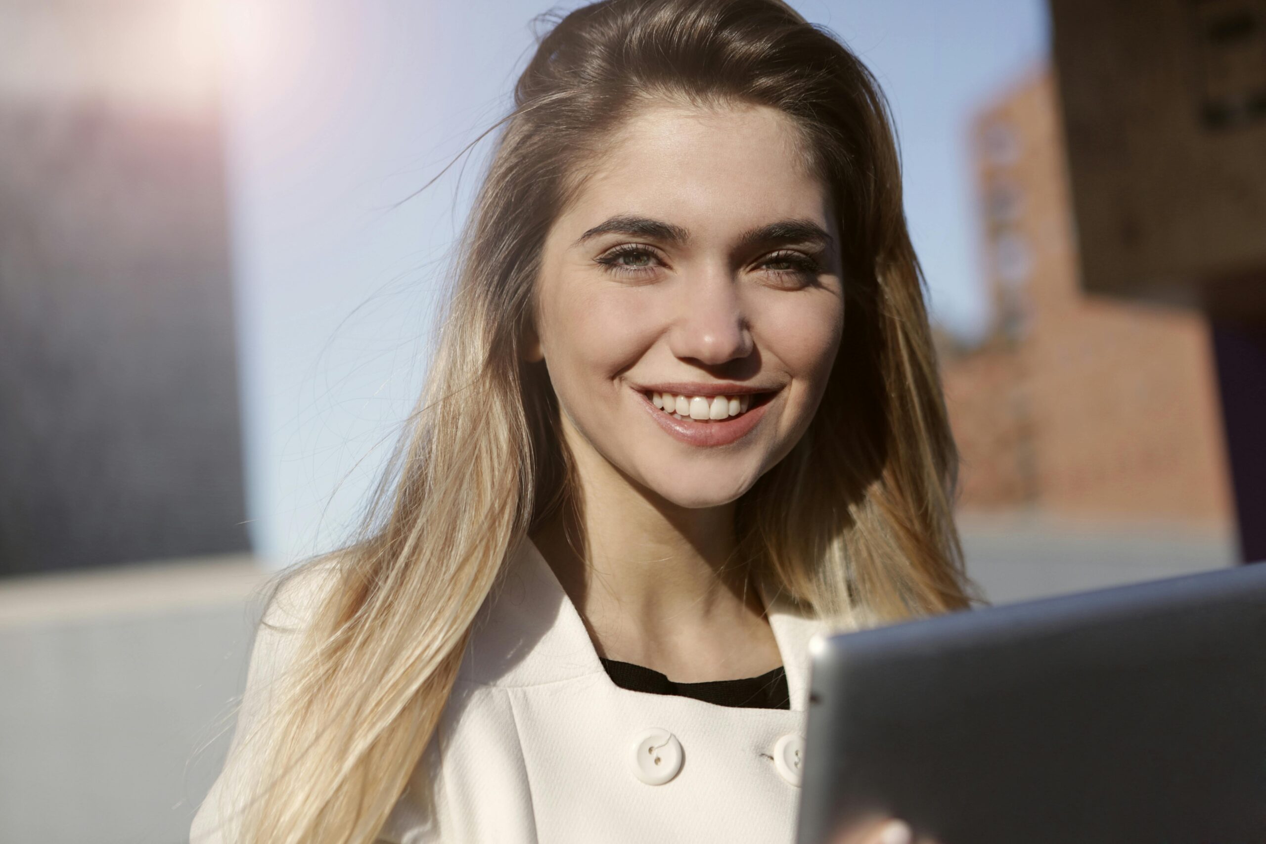 Portrait of a smiling blonde woman outdoors, holding a tablet and enjoying a sunny day.